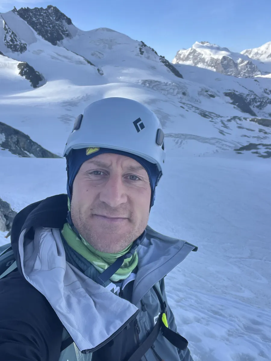 Thomas mit Helm und Schlauchtuch auf einem Gletscher, dahinter vergletscherte Gipfel